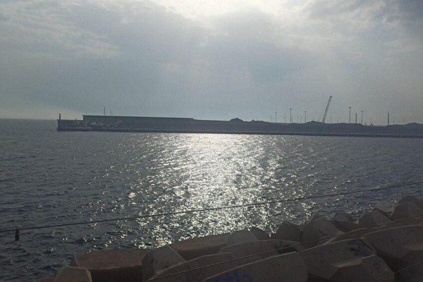 Een rustige zee met een groot schip in de verte, een golfbreker en industriële gebouwen aan de horizon. 3