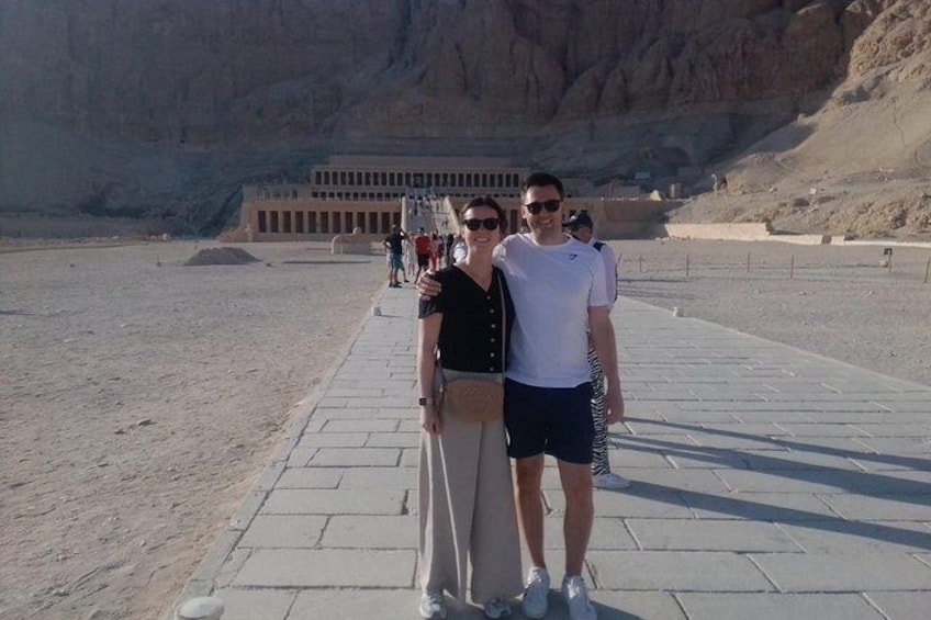 Two people standing on a paved walkway with a large rock formation and a building in the background. 4