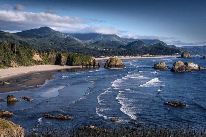 Un paisaje costero con una playa arenosa, afloramientos rocosos y montañas distantes bajo un cielo parcialmente nublado. 1