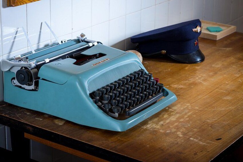 Een turquoise schrijfmachine op een houten tafel, met een blauwe militaire pet en een klein houten doosje op de achtergrond. 5