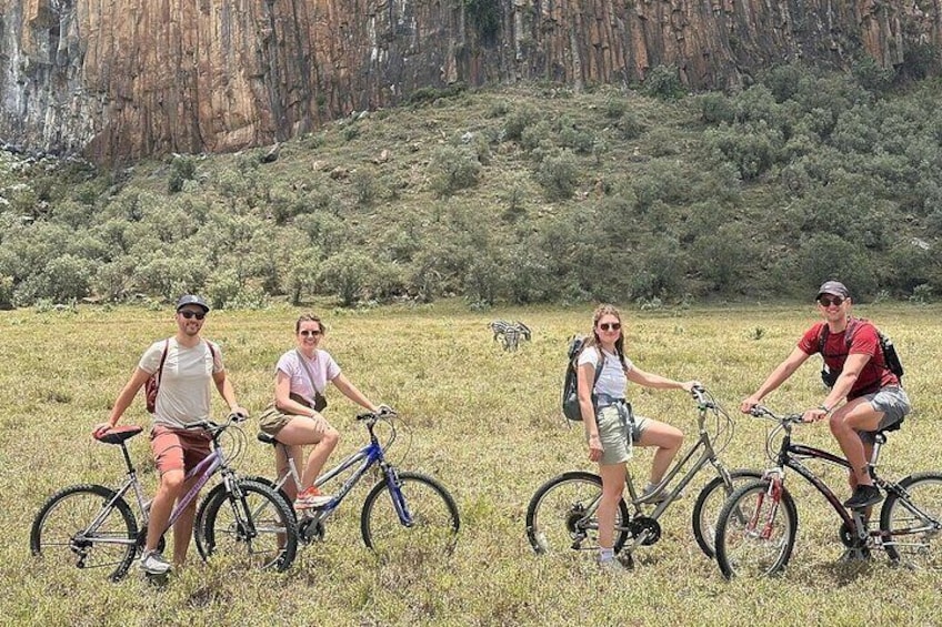 Four people on bicycles in a grassy field with a rocky hillside in the background. 1