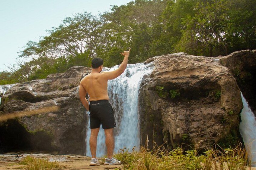 A man standing in front of a waterfall with his back to the camera, pointing at it. 5