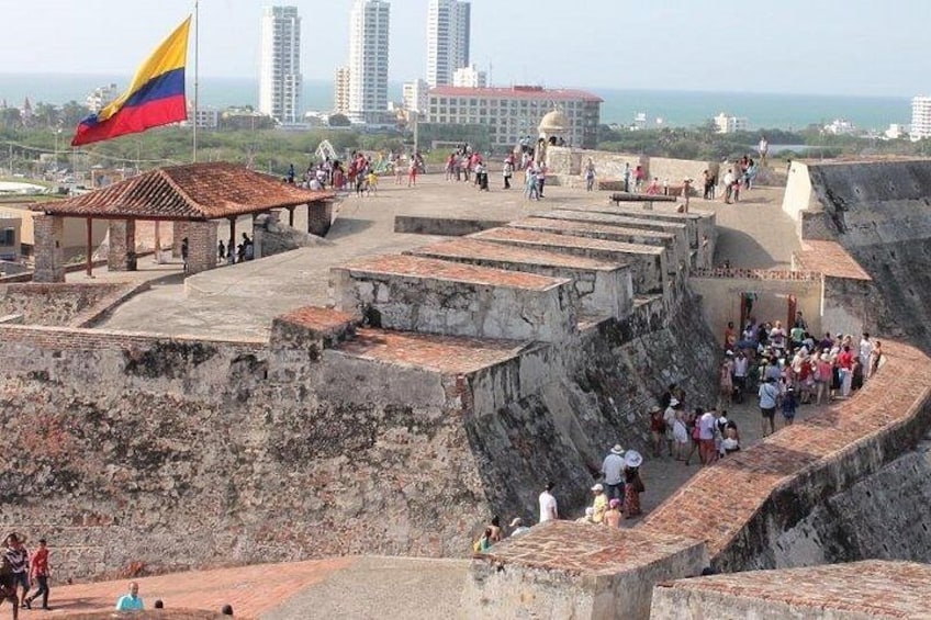 Eine historische Festung mit einer Flagge, Besuchern und modernen Gebäuden im Hintergrund. 1