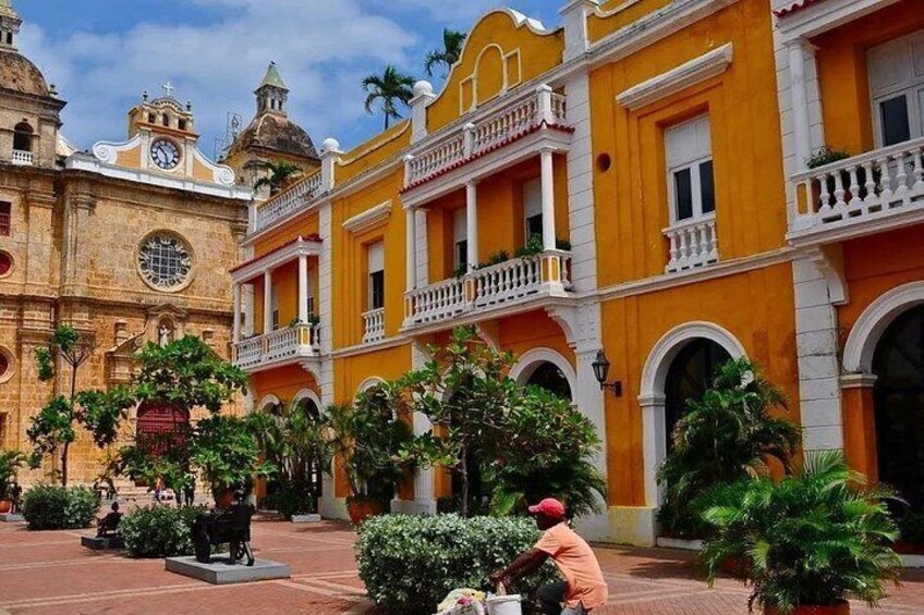 A colonial-style courtyard with buildings, palm trees, and a person riding a bicycle. 5