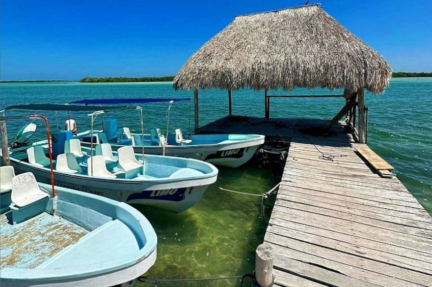 A wooden dock with two boats tied up, a thatched-roof structure, and clear turquoise water. 1