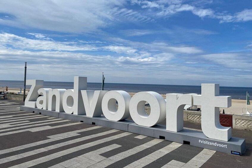 Ein großes weißes Schild mit dem Schriftzug 'Zandvoort' im Vordergrund, im Hintergrund ein Strand und Ozean. 5