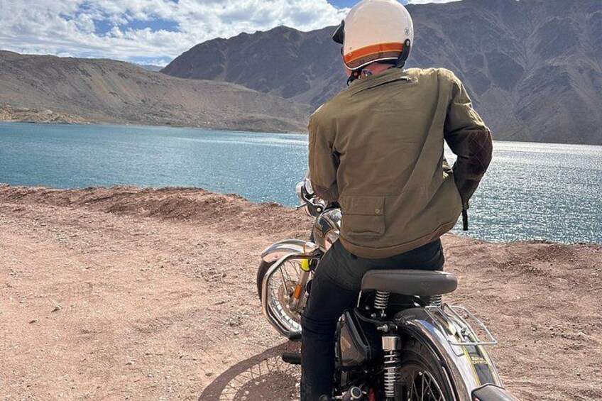 A person on a motorcycle by a lake with mountains in the background. 2