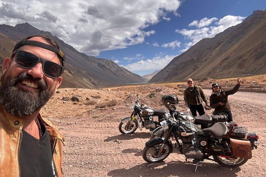 A desert landscape with mountains in the background, a person taking a selfie, and two motorcycles parked on the ground. 3
