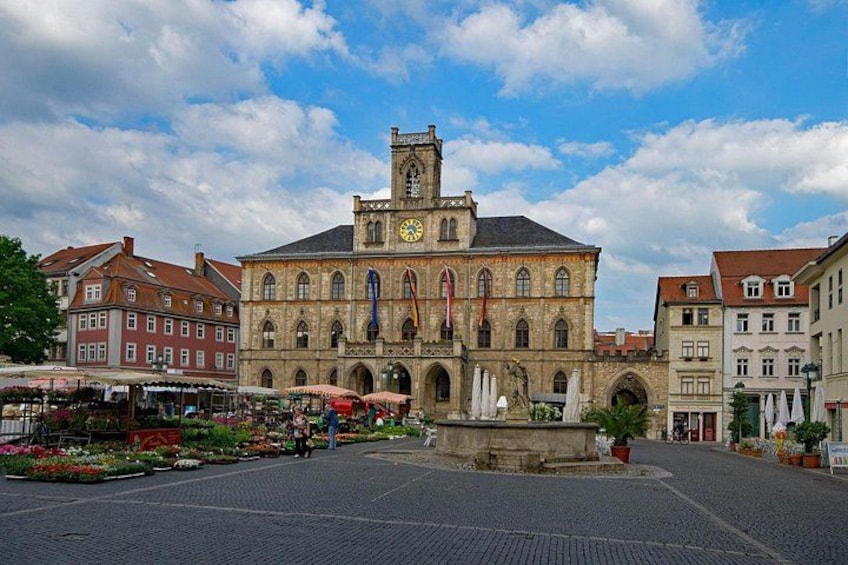 A historic building with a clock tower, surrounded by a cobblestone square with flower stalls. 1