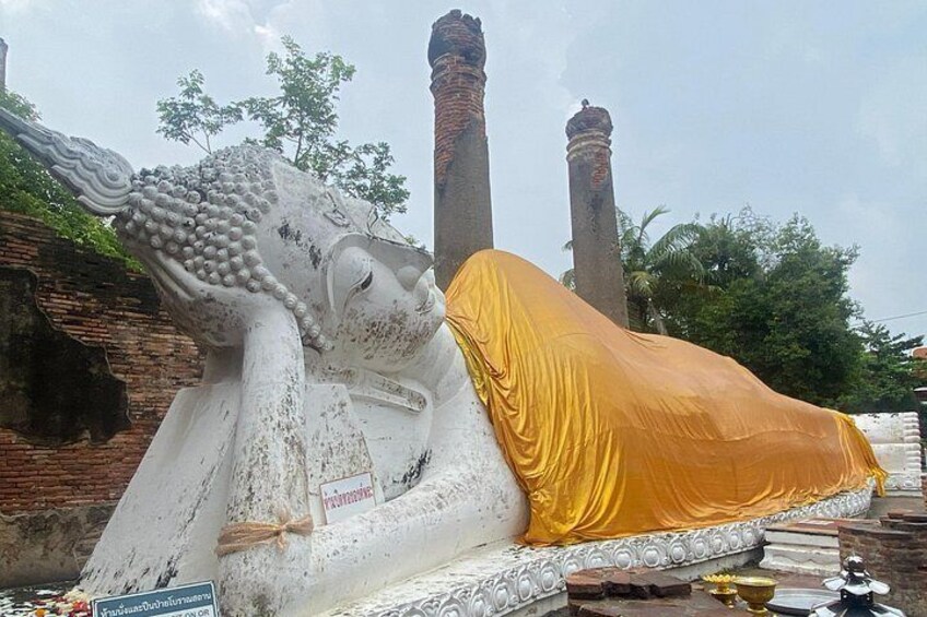 A large reclining Buddha statue covered with a yellow cloth, set against a backdrop of brick structures and trees. 1