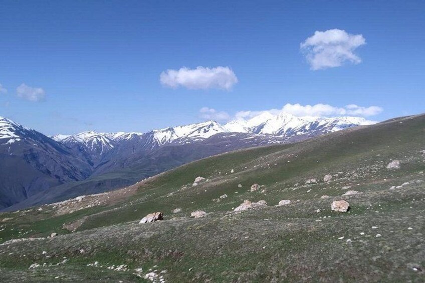 A mountainous landscape with snow-capped peaks in the distance, a clear sky, and scattered rocks on the foreground. 3