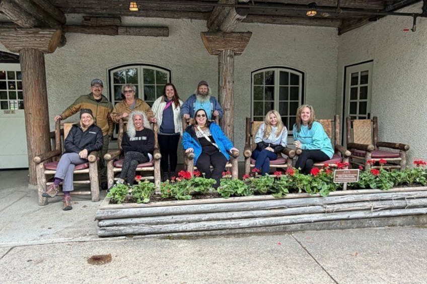 A group of people seated on wooden benches in a covered outdoor area with a stone floor, surrounded by potted plants and a stone wall. 4