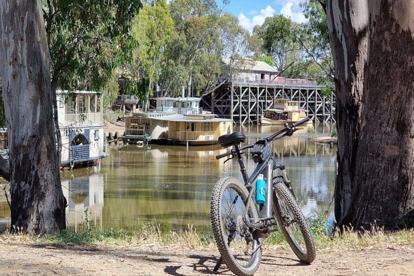 Una bicicleta de montaña estacionada junto a un río, con barcos y un puente al fondo. 4