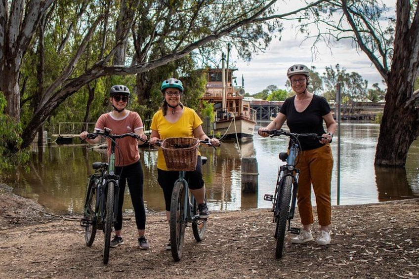 Tres personas en bicicletas cerca de un cuerpo de agua, con árboles y un puente al fondo. 1