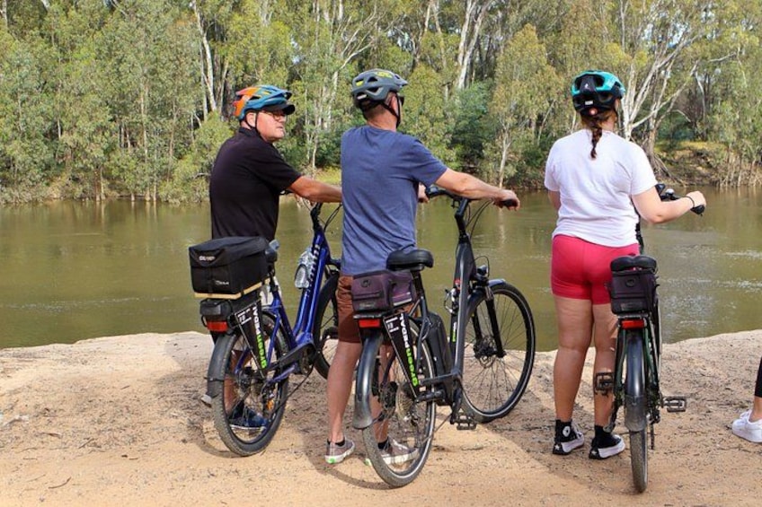 Tres personas en bicicletas a orillas de un río, con árboles al fondo. 3