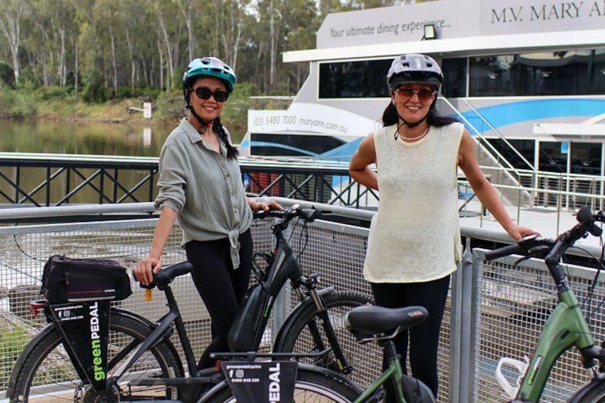 Dos personas de pie junto a bicicletas cerca de un cuerpo de agua con un bote al fondo. 2