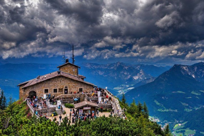 A mountain lodge with a steep staircase leading up to it, surrounded by lush greenery and a backdrop of rugged mountains. 1