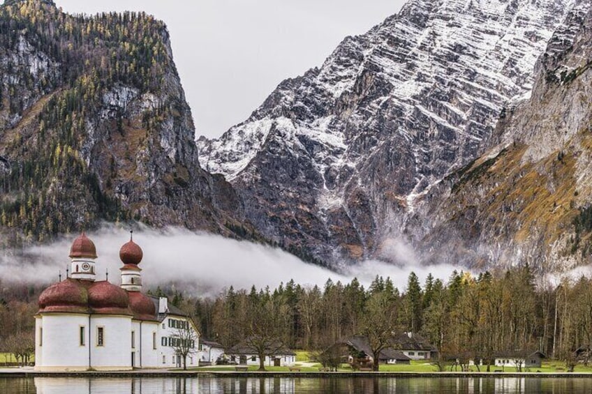 Una iglesia con cúpulas rojas junto a un lago, rodeada de montañas nevadas y colinas boscosas. 1