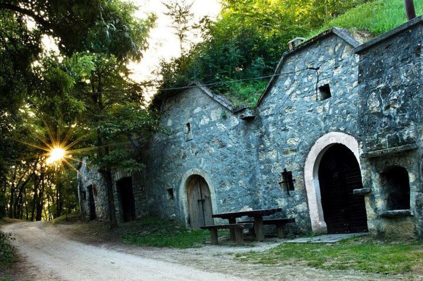 A rustic stone building with arched doorways, surrounded by trees and a dirt path. 2