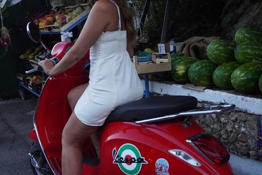 Una persona montando un scooter rojo con un vestido blanco, estacionado cerca de un puesto de frutas con sandías. 4
