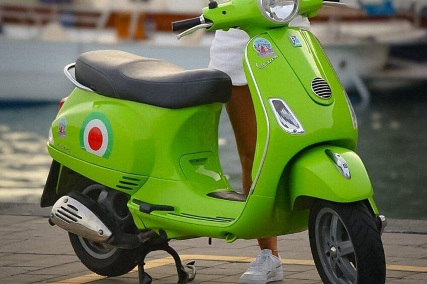 Un scooter verde brillante con asiento negro, un logo circular en el lateral y una persona de pie a su lado. 5
