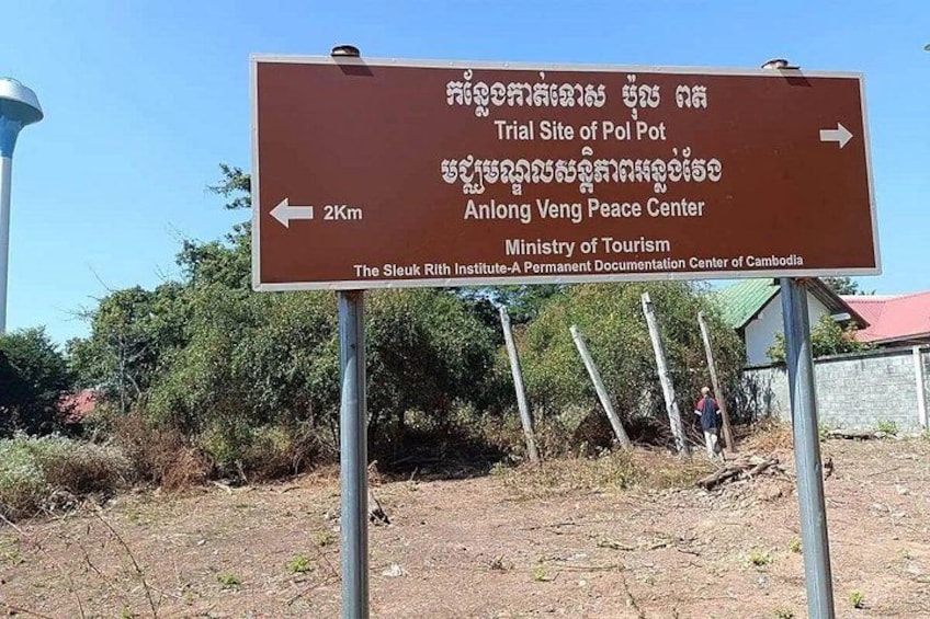 A directional sign with text in multiple languages, pointing towards the Trial Site of Pol Pot and the Anlong Veng Peace Center, managed by the Ministry of Tourism. 2