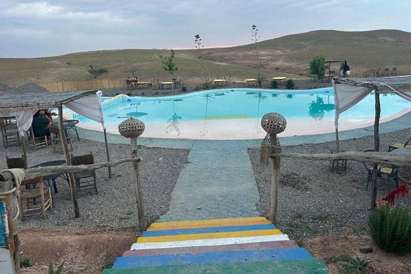 A swimming pool with a wooden deck, surrounded by tables and chairs, and a hilly landscape in the background. 5