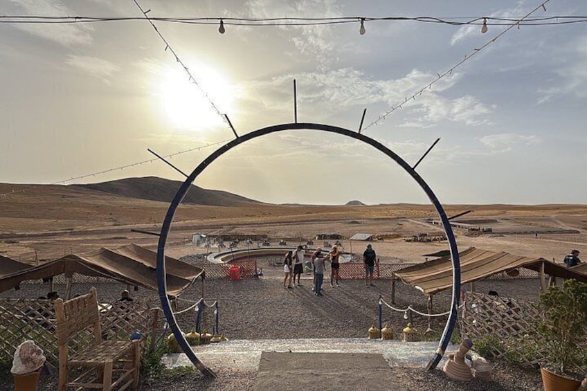 Paisaje desértico con un gran arco metálico, gente caminando y tiendas de campaña. 4