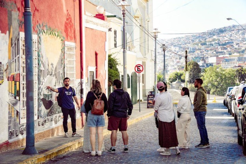 Un groupe de personnes marchant le long d’une rue pavée, une fresque murale sur un mur, des voitures garées et un paysage vallonné en arrière-plan. 2