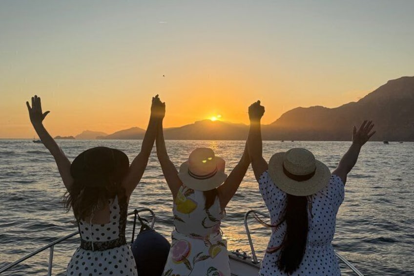 Three people on a boat with their arms raised, facing a sunset over water with mountains in the background. 1