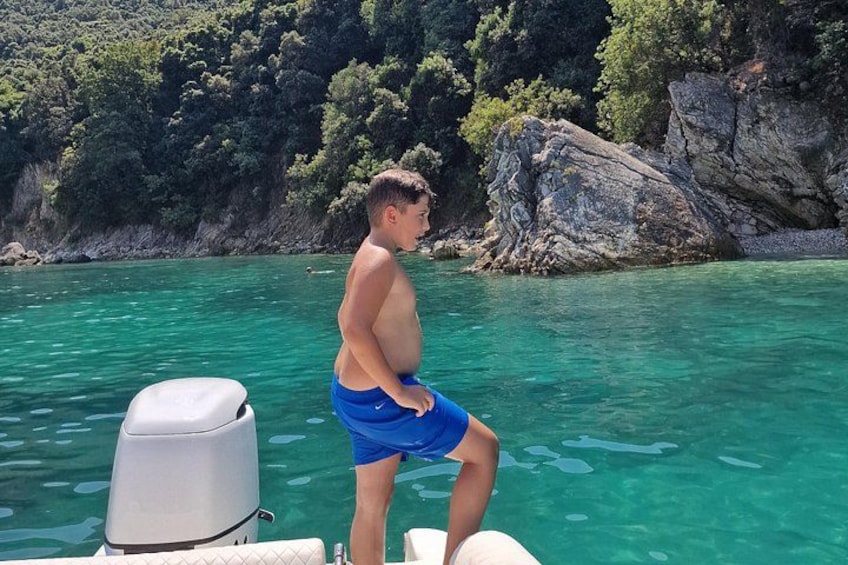 A person standing on a boat in clear water with a rocky shoreline and greenery in the background. 1