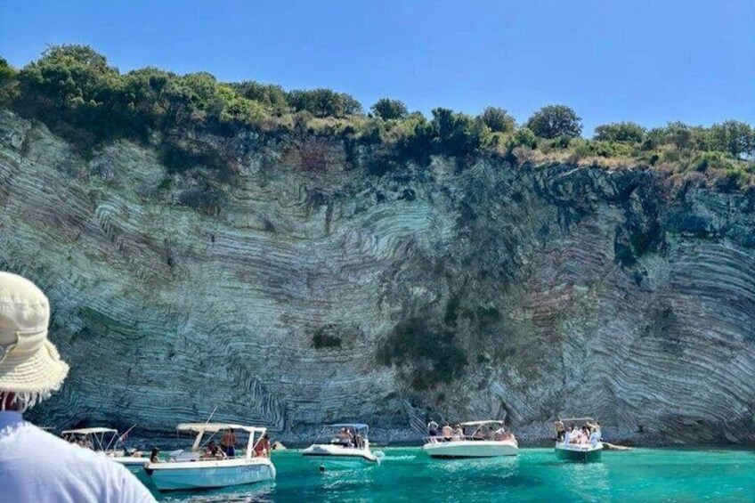 A coastal cliff with layered rock formations, boats on turquoise water, and a clear sky. 4
