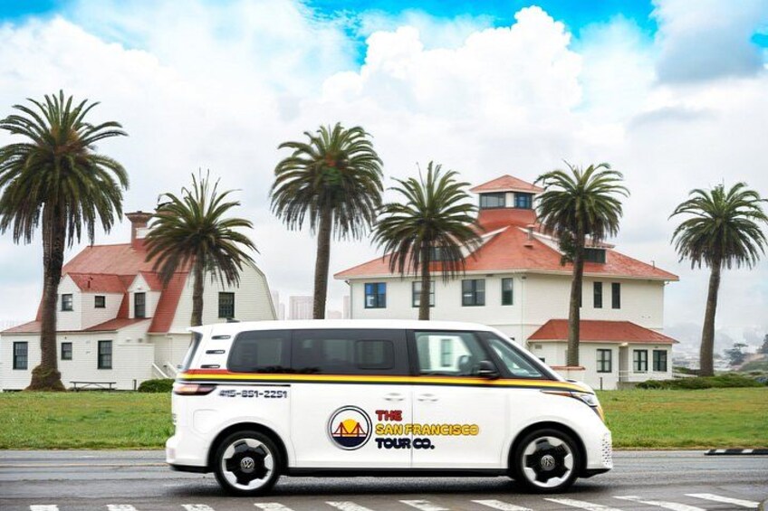 A white van with 'The Franciscan Tours Co.' logo parked in front of a large white building with a red roof, surrounded by palm trees. 3