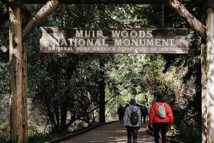 A wooden sign for Muir Woods National Monument with two hikers walking on a boardwalk. 1