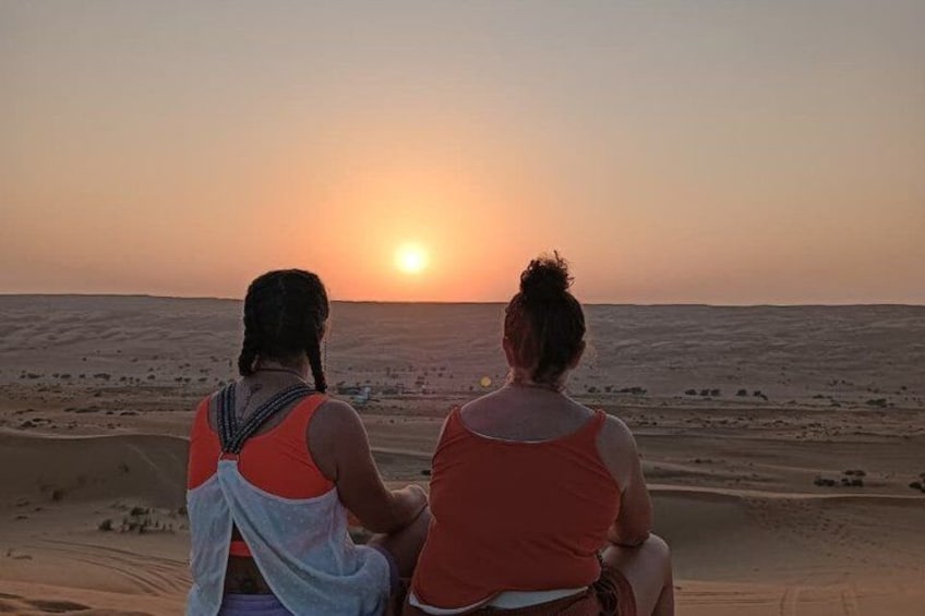 Dos personas sentadas en una duna, mirando una puesta de sol sobre un paisaje desértico. 3
