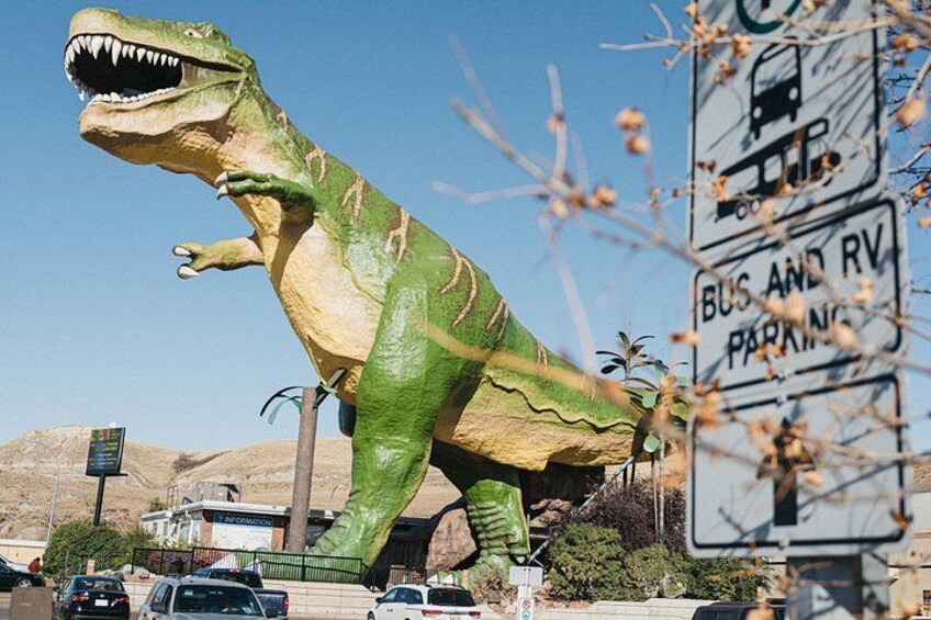 Een groot dinosaurusbeeld, een bord met parkeerplaatsen voor bus en campers, en een helderblauwe lucht. 2