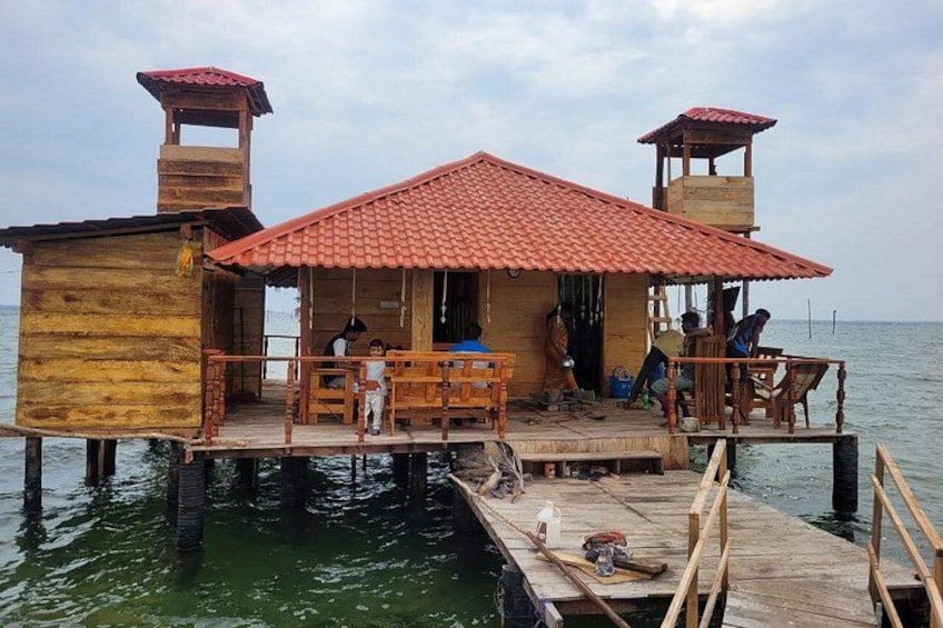 A wooden house with a red roof on stilts over water. 1