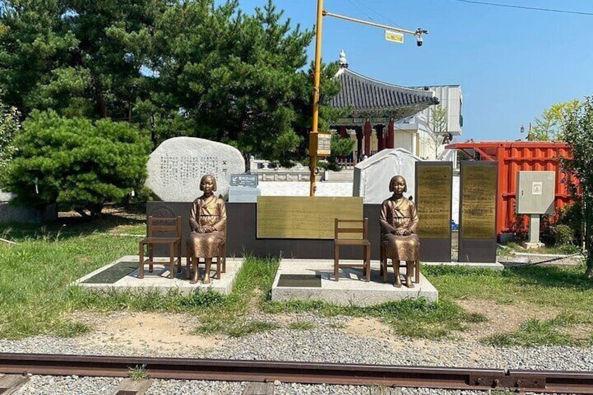 Two bronze statues of women seated on chairs, with a stone monument and a traditional pavilion in the background. 4