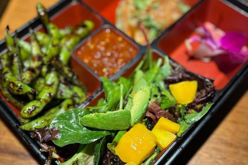 A meal with grilled vegetables, a salad, and mango pieces, served in a black dish with red sauce. 2