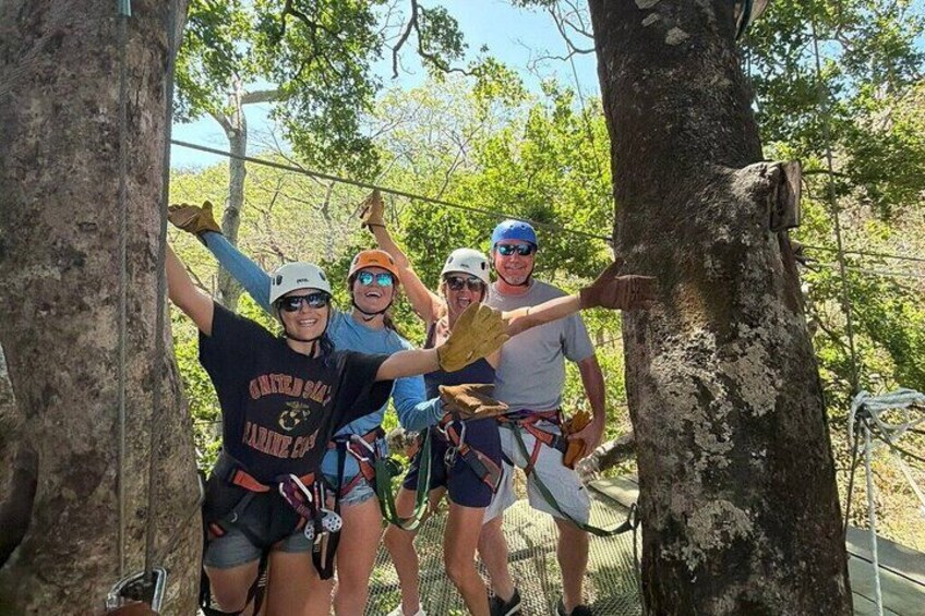 A group of people engaged in a zip-lining activity, wearing helmets and harnesses, with trees and a clear sky in the background. 5