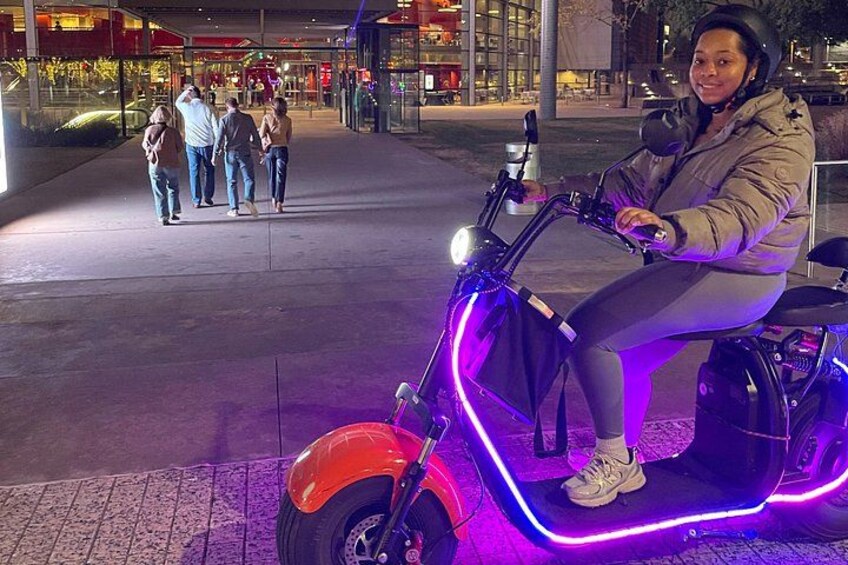 Een persoon rijdt op een scooter met neonverlichting, een geplaveid voetpad en een groep mensen op de achtergrond. 1
