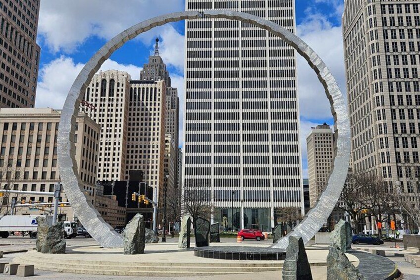 Une vue urbaine avec une grande sculpture circulaire au premier plan, de hauts immeubles en arrière-plan et une voiture rouge garée près de la sculpture. 1
