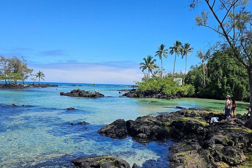 A clear blue sea with rocks, palm trees, and a person standing on the rocks. 2