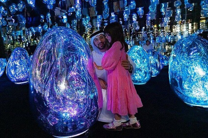 A person in traditional attire embracing a child in a pink dress in a setting with large illuminated spheres. 1
