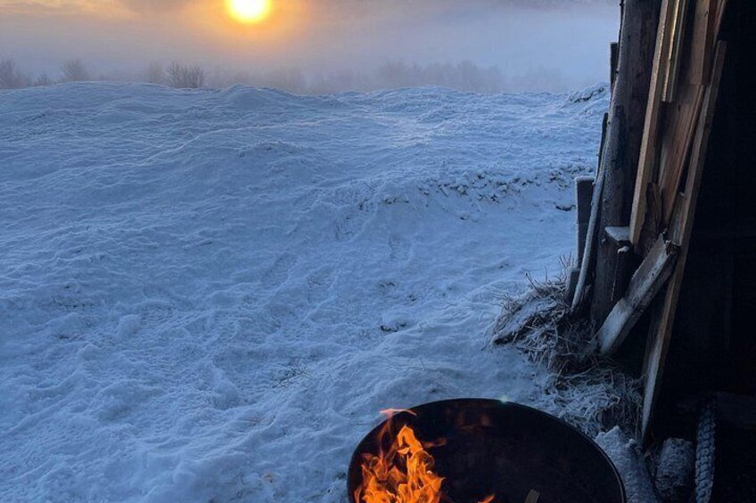 A snowy landscape with a fire pit and a wooden structure. 1