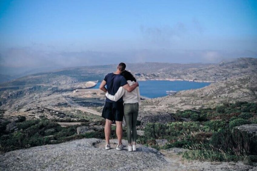 Dos personas abrazándose sobre un promontorio rocoso con vistas a un paisaje costero, con una masa de agua y colinas distantes. 1
