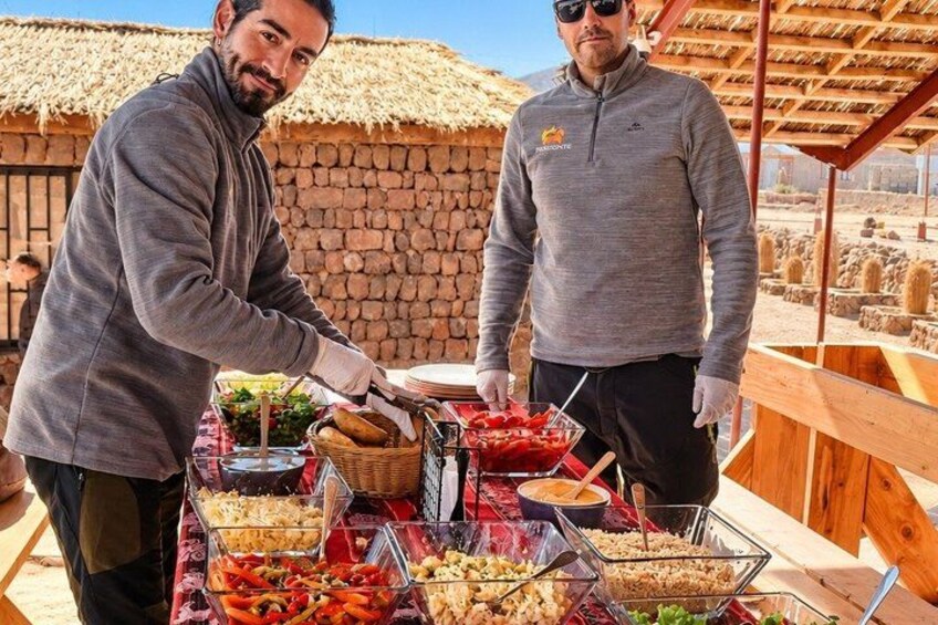 Dos personas sirven comida en un bufé al aire libre, con diversos platos y una estructura de techo de paja al fondo. 3