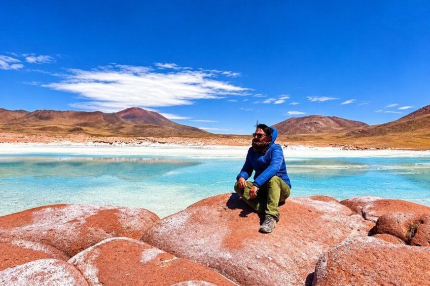 Una persona sentada sobre rocas rojas junto a un lago azul cristalino, con montañas al fondo. 5