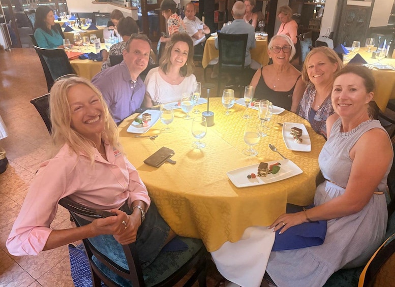 A group of people seated around a round table with yellow tablecloths, enjoying a meal with various dishes and glasses of wine. 2