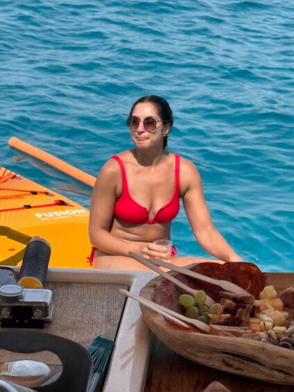 A person in a red bikini sitting on a boat with a paddleboard nearby, surrounded by water. 1